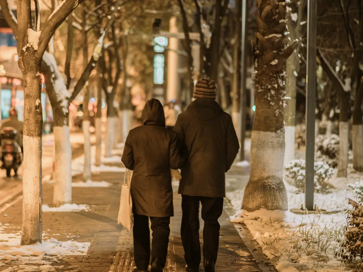 Un couple, de dos, marche bras dessus bras dessous sur une allée enneigée éclairée.