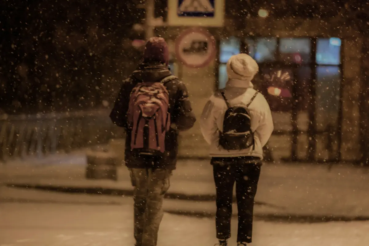 Deux amis de dos, sacs au dos, marchent sous la neige qui tombe un soir d'hiver.