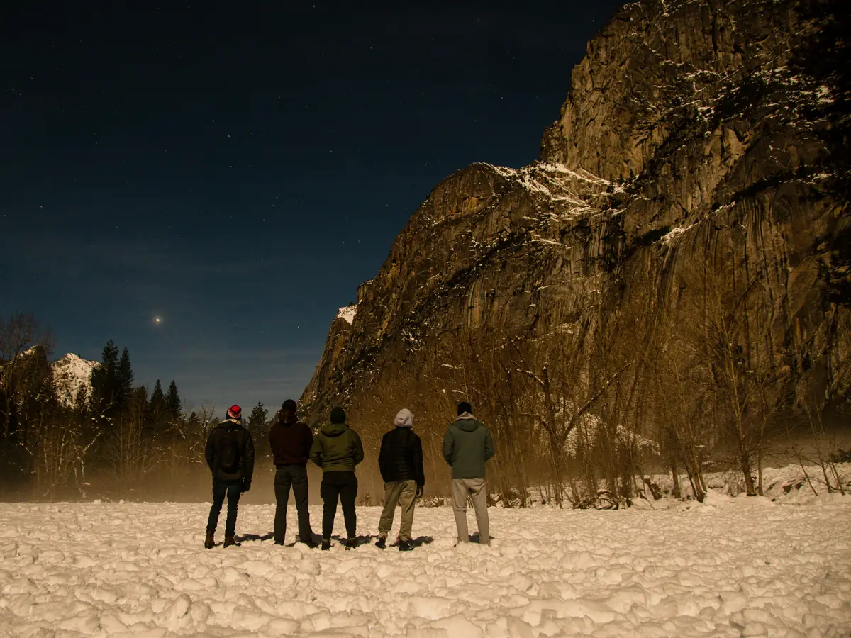 Un groupe de cinq personnes, de dos, contemple une falaise et le ciel étoilé dans la neige.