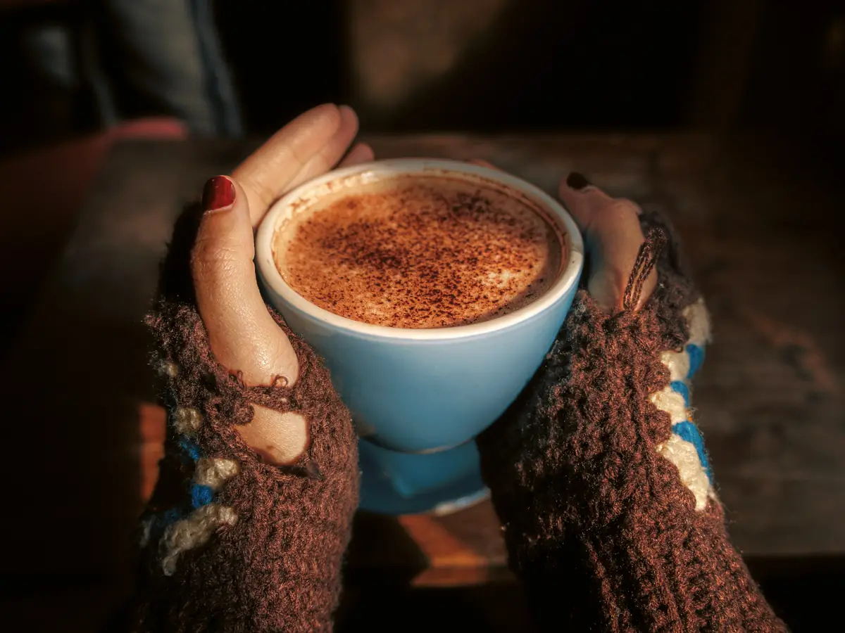Des mains en mitaines sans doigts entourent une tasse de chocolat chaud.