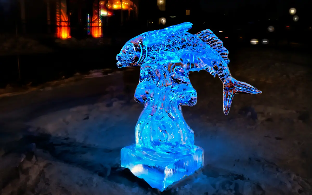 Sculpture de glace en forme de poisson, éclairée en bleu, la nuit dans la neige.