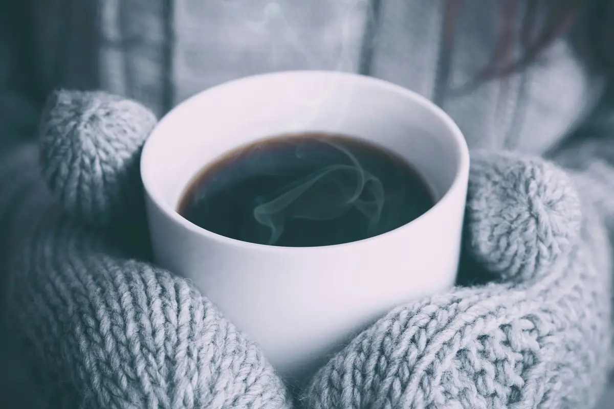 Des mitaines en tricot gris tiennent une tasse de café fumant.