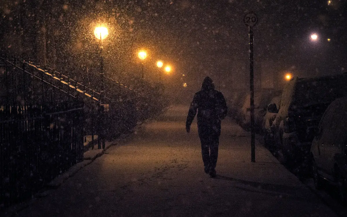 Un passant marche sur un trottoir enneigé sous des lampadaires orangés pendant une chute de neige.