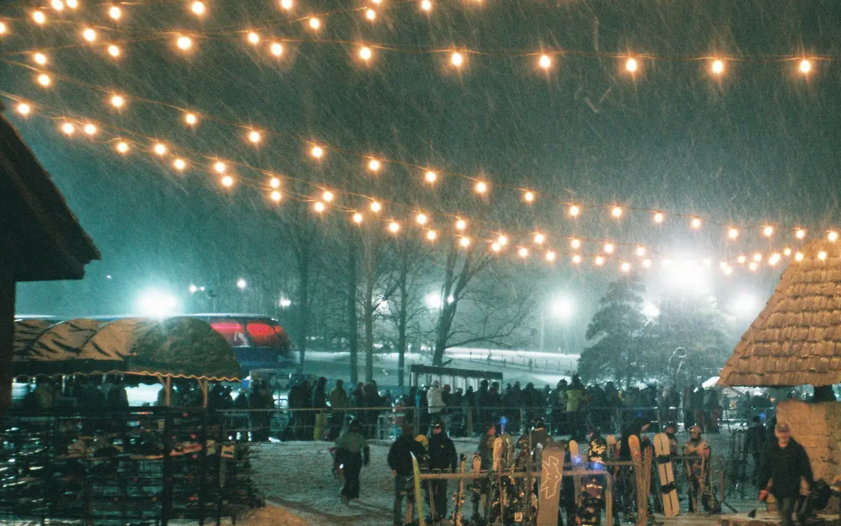 String lights over a busy snowy plaza during heavy evening snowfall.